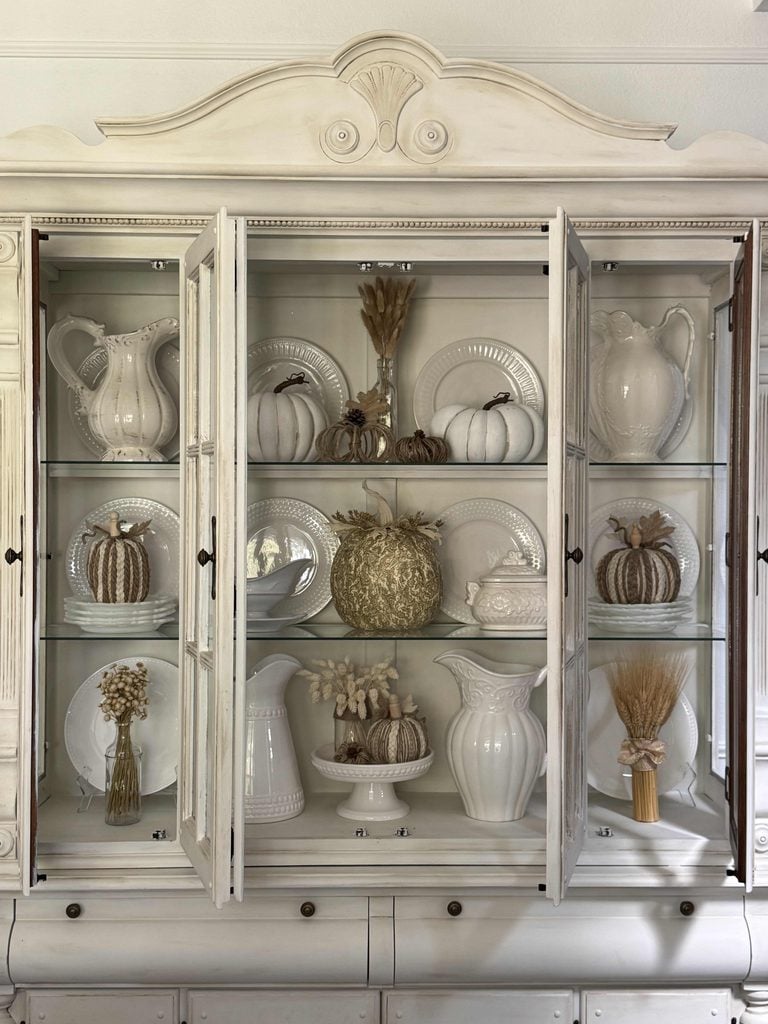 A glass-front cabinet displays white pitchers, plates, and vases, along with decorative pumpkins and dried floral arrangements in a neutral, rustic style. The items are neatly arranged on three shelves.