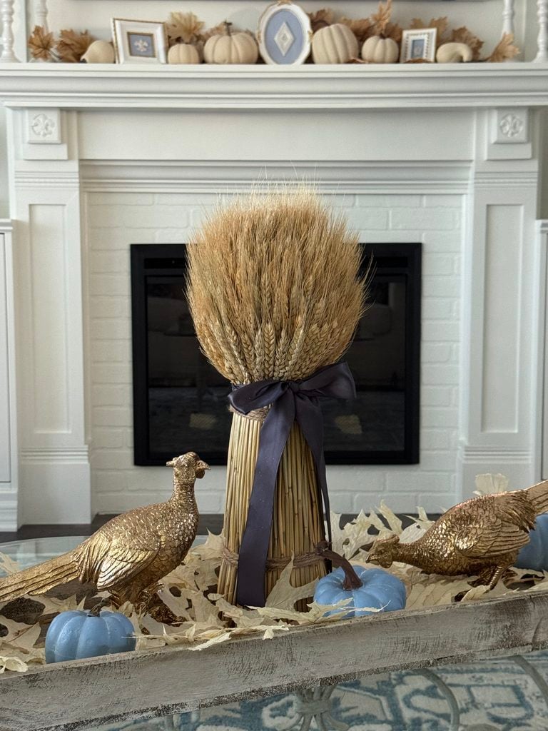 A bundle of wheat tied with a ribbon is centered between two gold pheasant figurines and blue pumpkins, sitting on a tray in front of a white fireplace decorated with neutral pumpkins and framed art.
