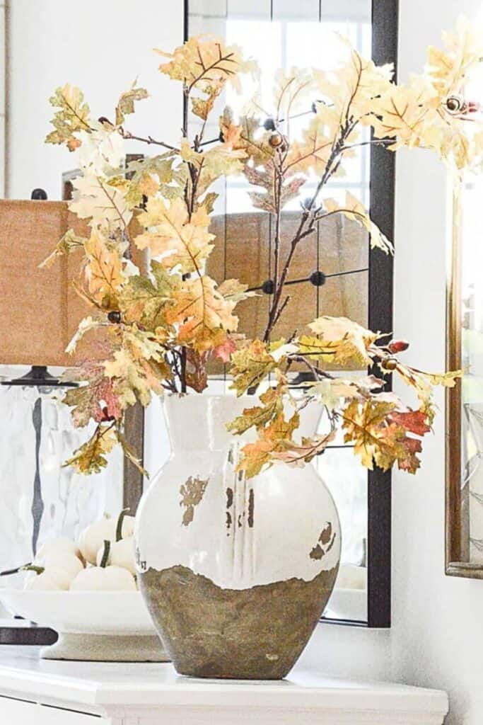 A ceramic vase with brown and white glaze holds branches of autumn leaves in yellow, orange, and red tones. The arrangement sits on a white surface in front of a mirror and next to a lamp.