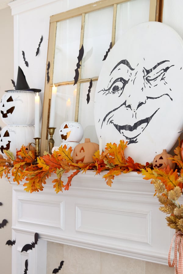 A festive mantle decorated with autumn leaves, white and orange pumpkins, gold candlesticks, paper bats, and a large oval sign featuring a winking moon face.