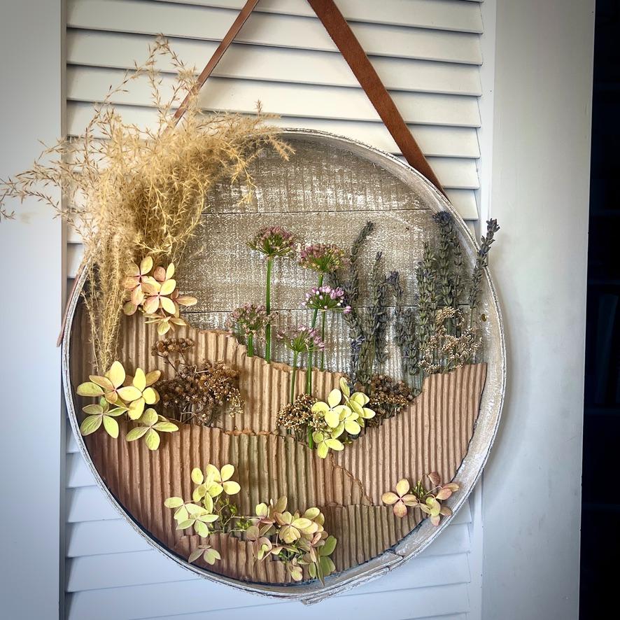 A round, shallow frame hangs by a brown strap, filled with dried flowers and grasses arranged on corrugated cardboard in an artistic display set against a white shuttered background.