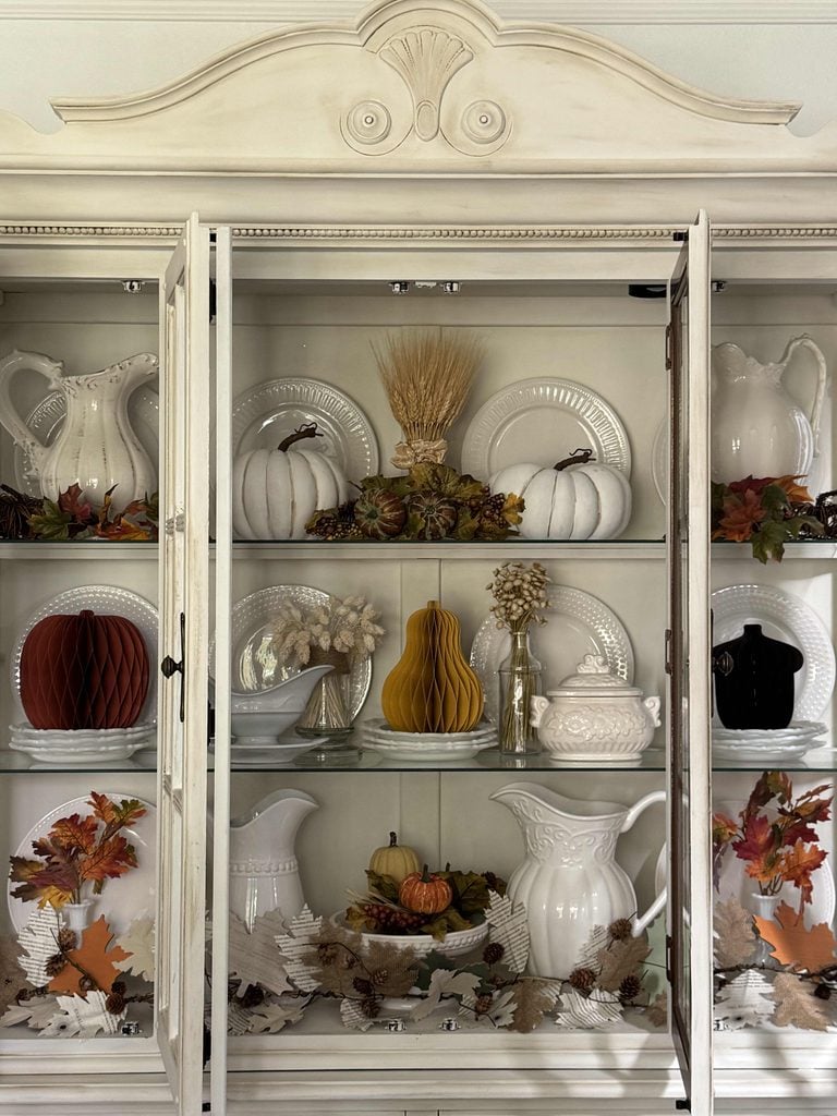 A cream china cabinet displays white dishes, pitchers, pumpkins, paper decorations, and autumn leaves, creating a cozy fall-themed arrangement behind glass doors.