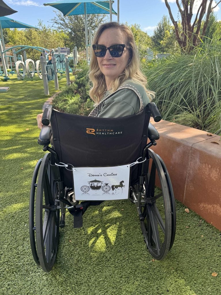 A woman with long blonde hair and sunglasses sits outside in a wheelchair on green grass, facing away from the camera. Plants, playground equipment, and blue sunshades are visible under a sunny sky.