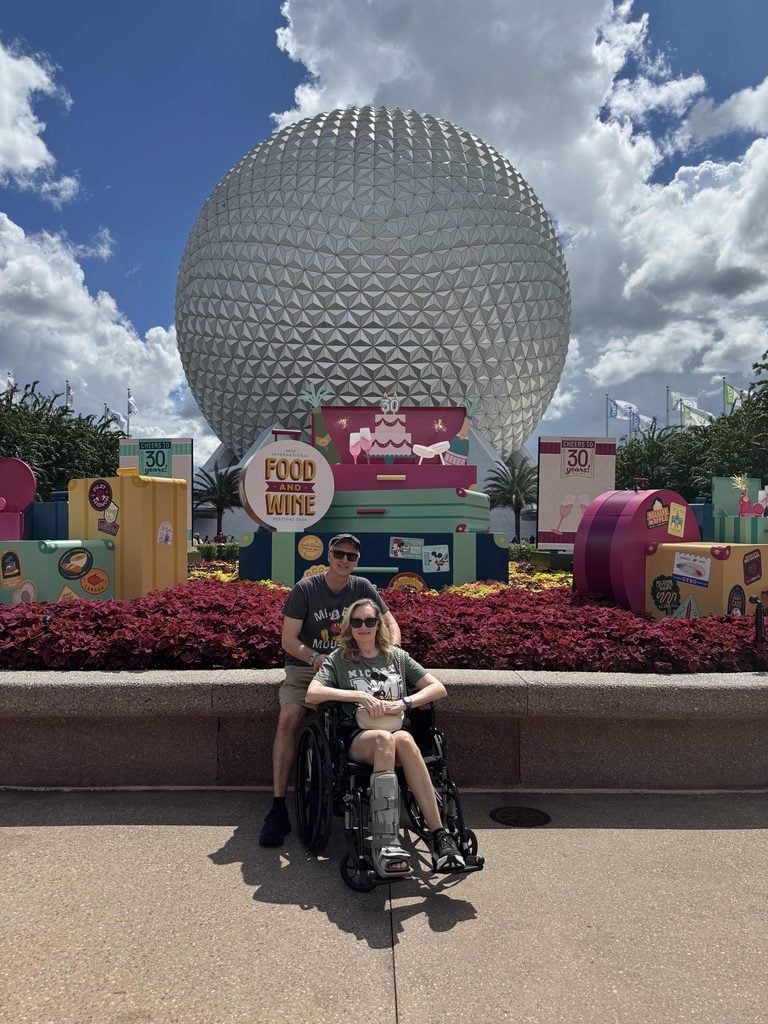 A man stands behind a woman in a wheelchair in front of Epcot’s Spaceship Earth at Disney World, surrounded by colorful Food & Wine Festival decorations on a sunny day.