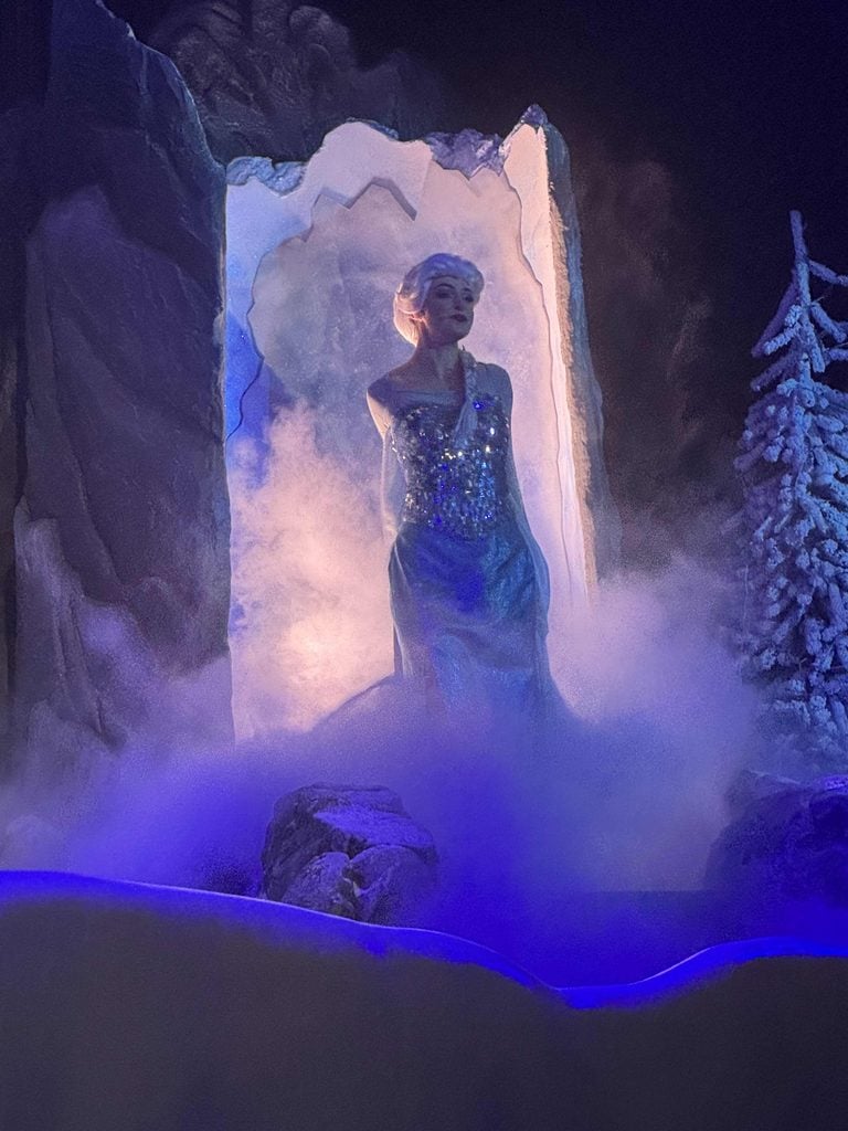 A person in a sparkling blue dress and white wig stands in front of an icy archway, surrounded by mist and artificial snow, with a frosty tree and rocks nearby, all illuminated by blue and purple lights.