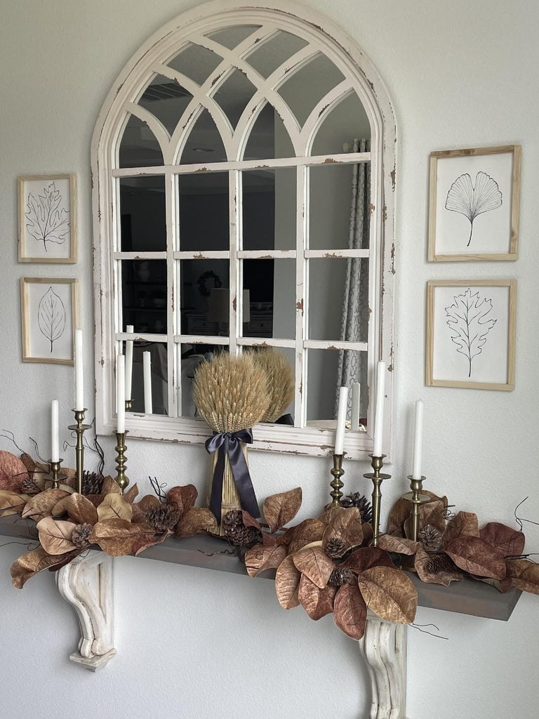 A mantel decorated with brass candlesticks, brown dried leaves, pinecones, and a wheat bouquet in front of a vintage white arched mirror, with four framed botanical sketches on the wall.