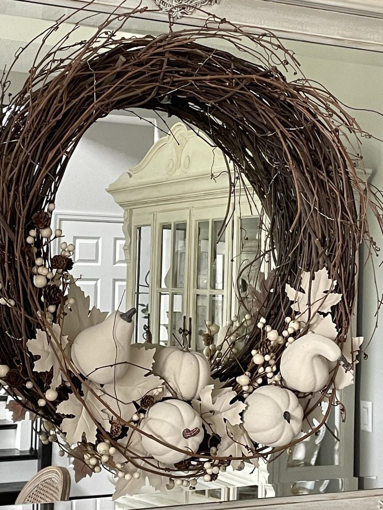 A fall wreath made of twigs, decorated with white pumpkins, white berries, and cream-colored leaves, hangs indoors in front of a mirrored cabinet.