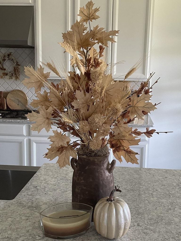 A brown vase with dried autumn leaves and wheat sits on a kitchen counter beside a white pumpkin and a large beige candle in a glass container, creating one of the simplest natural fall decor ideas for a cozy seasonal touch.