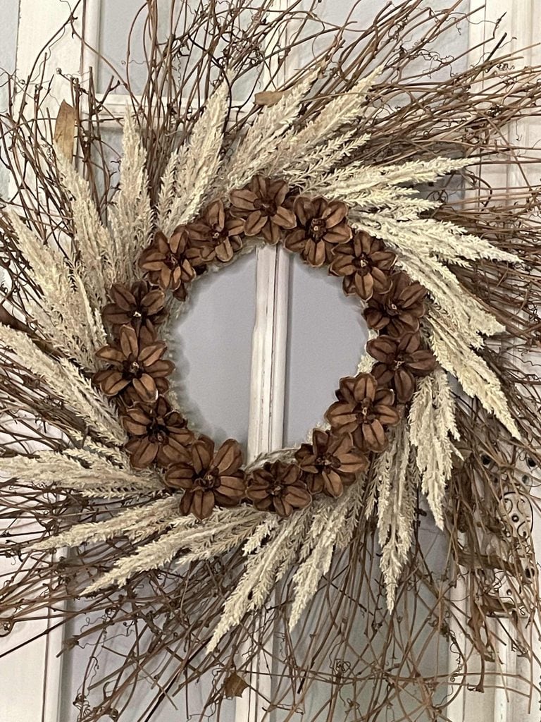 A decorative wreath made of dried brown flowers, beige grasses, and twigs hangs on a white paneled door, evoking the rustic charm of a DIY Twig Pumpkin centerpiece.