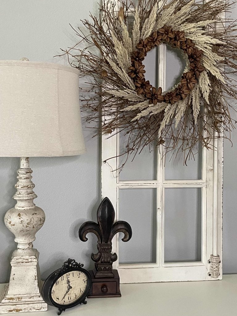 A rustic white lamp, vintage clock, fleur-de-lis statue, and a dried wreath on an old white window frame are arranged on a white tabletop against a light gray wall.