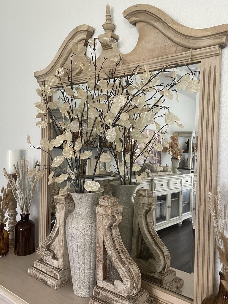 A beige vase with cream-colored, decorative branches sits on a mantle in front of a wooden-framed mirror. The mirror reflects a sideboard and more neutral-toned decor in the background.