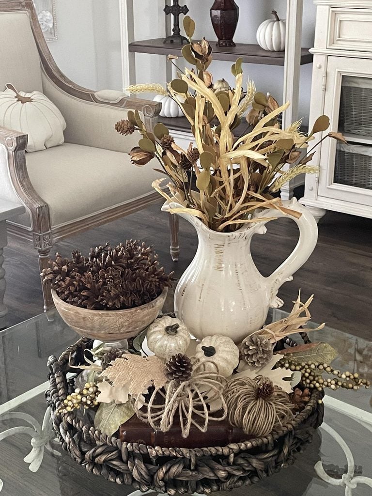 A fall-themed centerpiece with a white pitcher holding dried foliage, a bowl of pinecones, decorative pumpkins, and twine balls arranged on a woven tray on a glass table in a cozy living room.