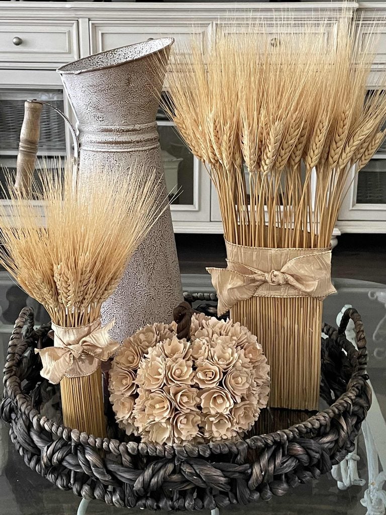 A decorative tray holds wheat bundles tied with ribbon, a rustic metal pitcher, and a beige flower-covered pumpkin alongside a charming DIY Twig Pumpkin, set on a glass table with white cabinets in the background.