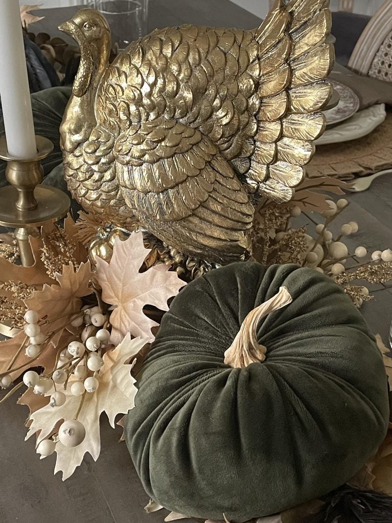 A gold turkey figurine sits among dried leaves, white berries, and a green velvet pumpkin on a decorated table, evoking an autumn or Thanksgiving theme.