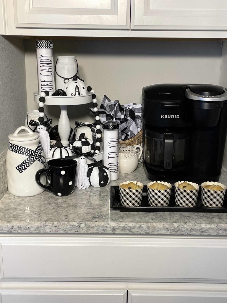A black Keurig coffee maker sits on a countertop with black and white mugs, buffalo plaid pumpkins, a milk jug, jars, and four black-and-white checkered muffins, all in a black and white themed coffee station decor.