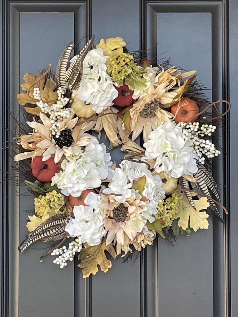 A fall wreath with white flowers, cream sunflowers, small pumpkins, faux berries, leaves, and striped feathers hangs on a dark paneled door.