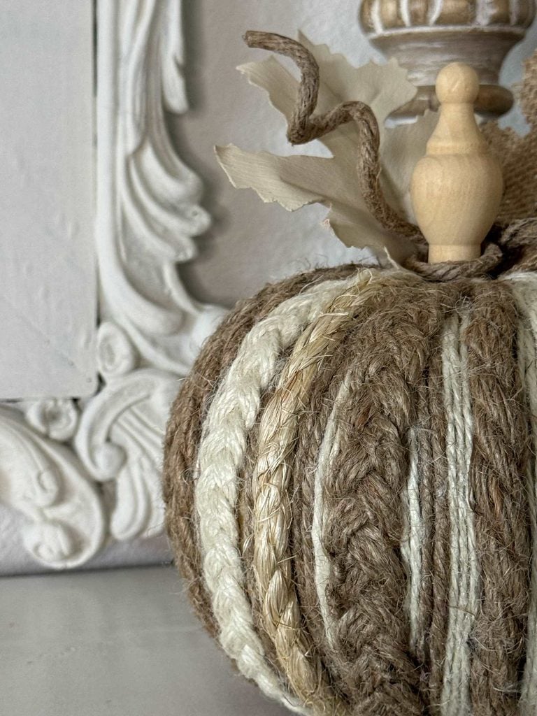 A decorative pumpkin made of braided twine and cream rope, topped with a wooden stem and fabric leaves, sits on a light surface in front of an ornate white frame.