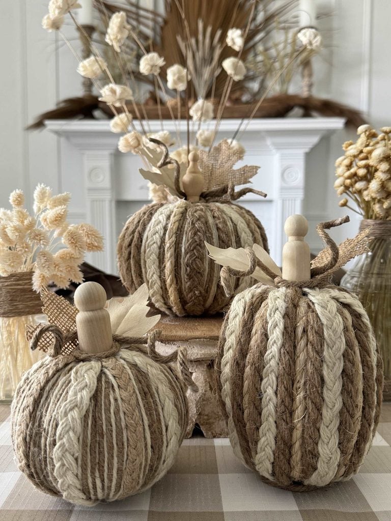 Three decorative pumpkins made from braided jute and cream rope, each topped with a wooden peg stem, sit on a plaid cloth with dried floral arrangements in the background.