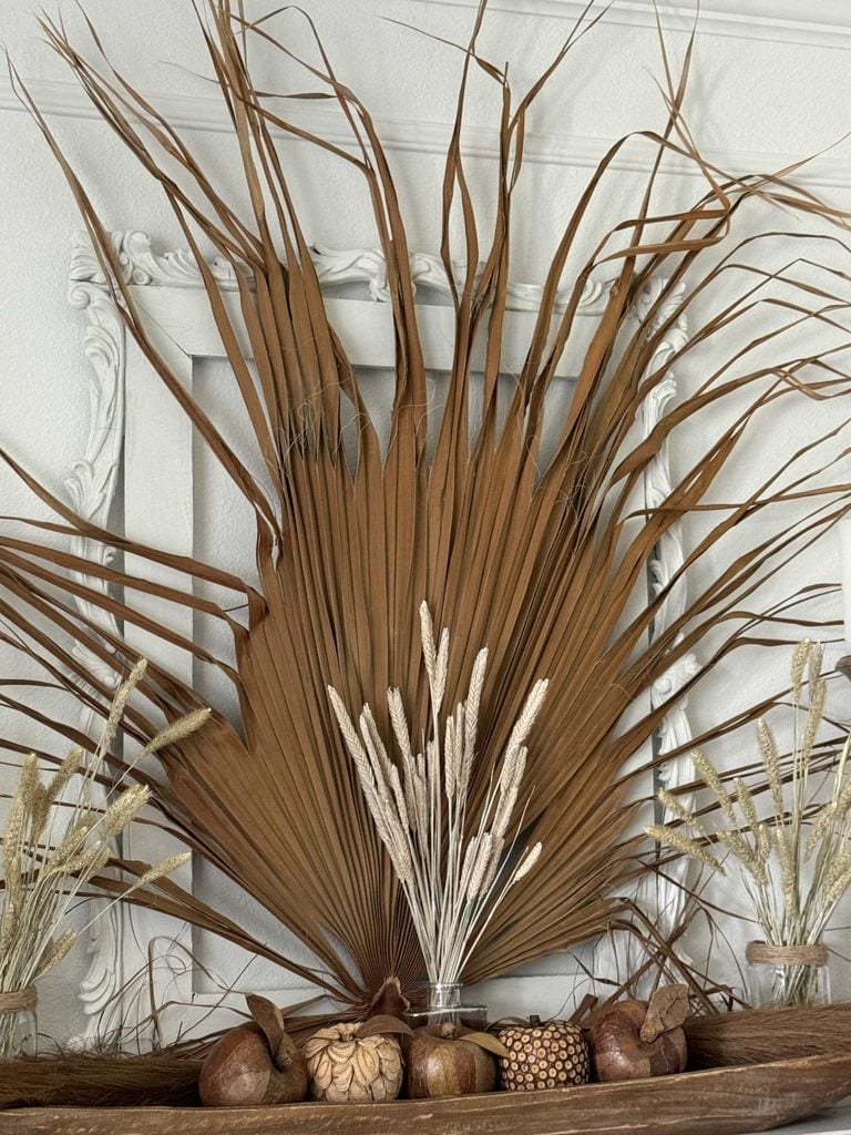 Dried palm leaves and grasses arranged in glass jars sit on a wooden shelf, with decorative gourds and seed pods in front. A white ornate frame leans against a light wall in the background.