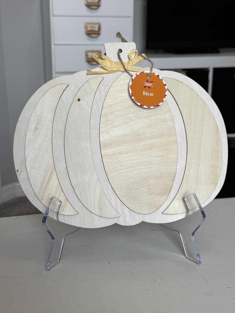 A blank wooden pumpkin-shaped decor piece sits on a clear stand, with an orange tag labeled Décor and a raffia bow on top, set on a light-colored table indoors.