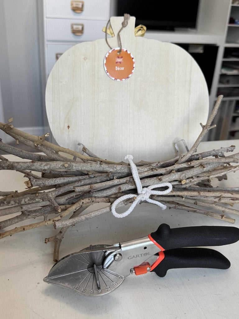 A blank wooden sign, a bundle of sticks tied with white rope, and a pair of pruning shears on a white surface set the stage for your next creative project—with shelves and storage drawers providing an organized backdrop.