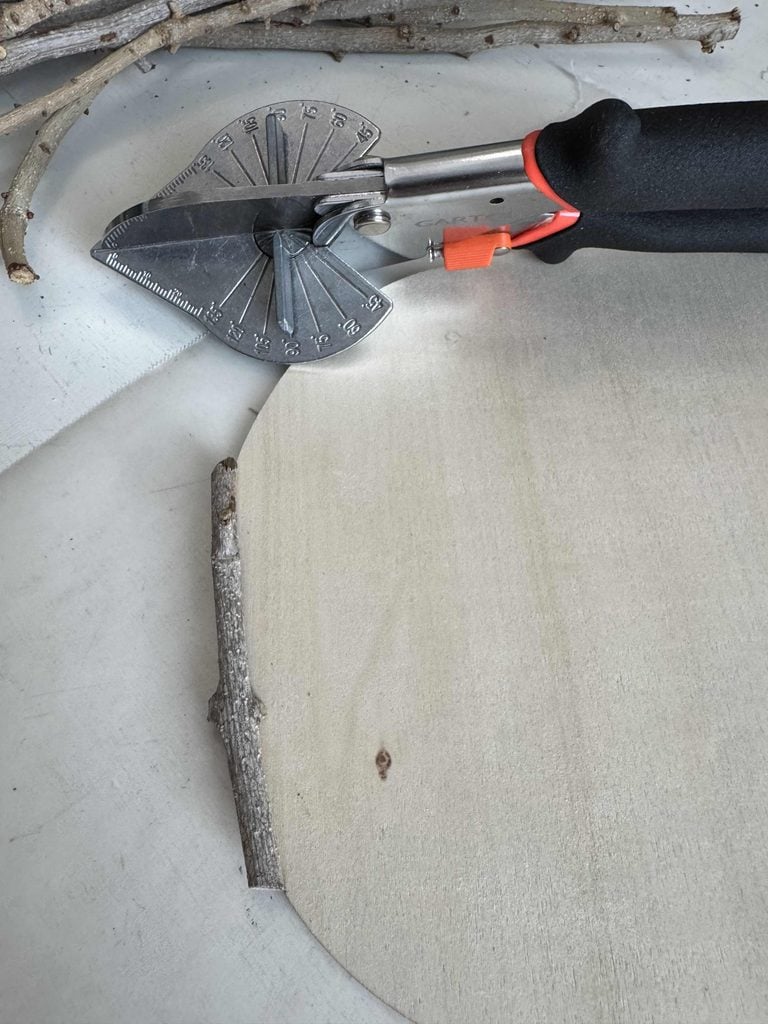 A miter shear tool with an orange and black handle rests on a light wood surface next to a small branch and several twigs. The tool’s angled blade and measurement markings are clearly visible.