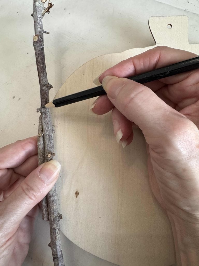 A person holds a twig against a wooden shape and marks it with a pencil, preparing the twig for cutting.
