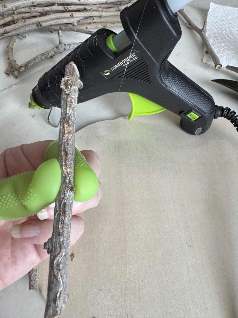 A hand with two green finger protectors holds a stick near a hot glue gun, with several sticks and a paper towel in the background on a light surface.