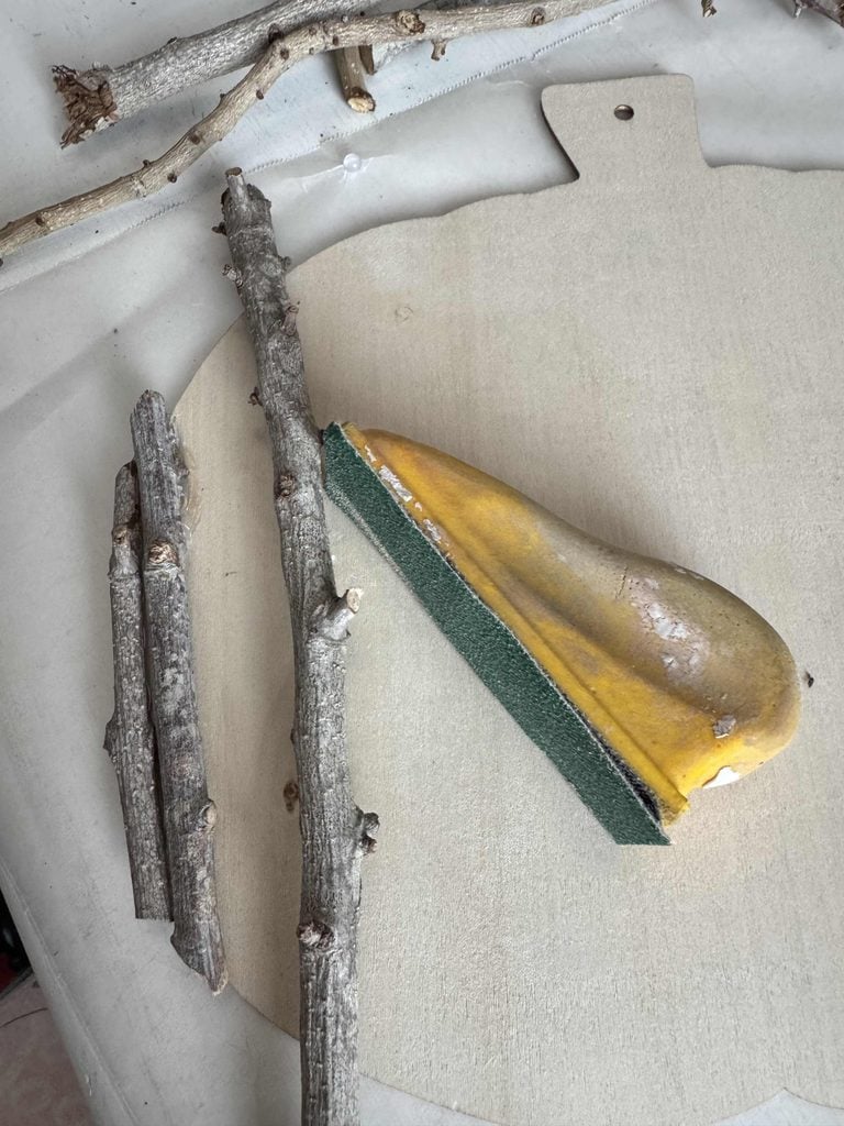 A yellow ceramic pear with a green sponge base sits on a light wooden cutting board next to three rough tree branches, creating a charming scene reminiscent of a DIY Twig Pumpkin craft.