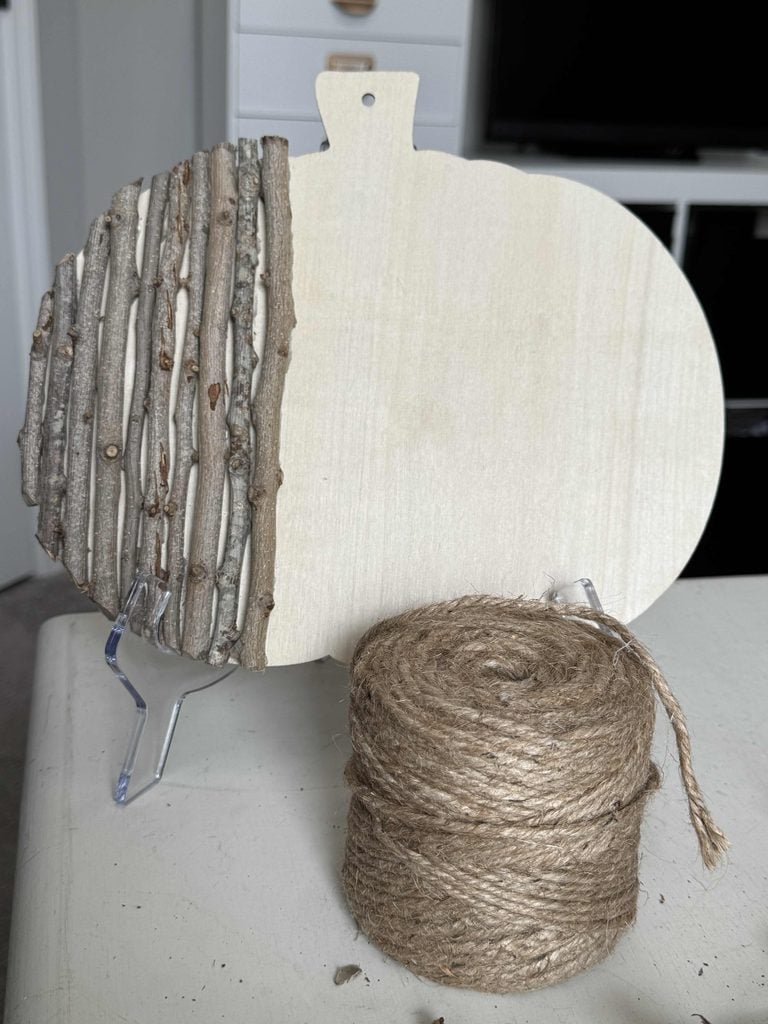 A wooden pumpkin-shaped sign on a stand, half decorated with vertical sticks in the style of a DIY Twig Pumpkin. A spool of twine sits in front on a table, while drawers and a TV are in the background.