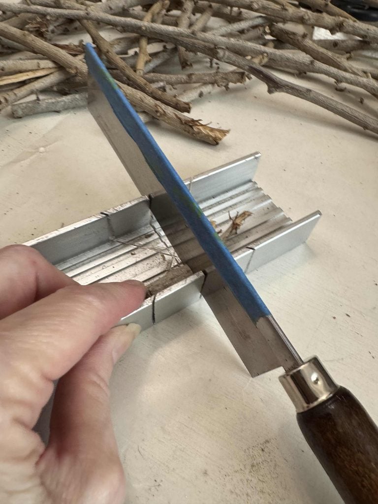 A hand uses a saw and a miter box to cut a small stick, preparing materials for a DIY Twig Pumpkin. Several more sticks are scattered on the surface in the background.