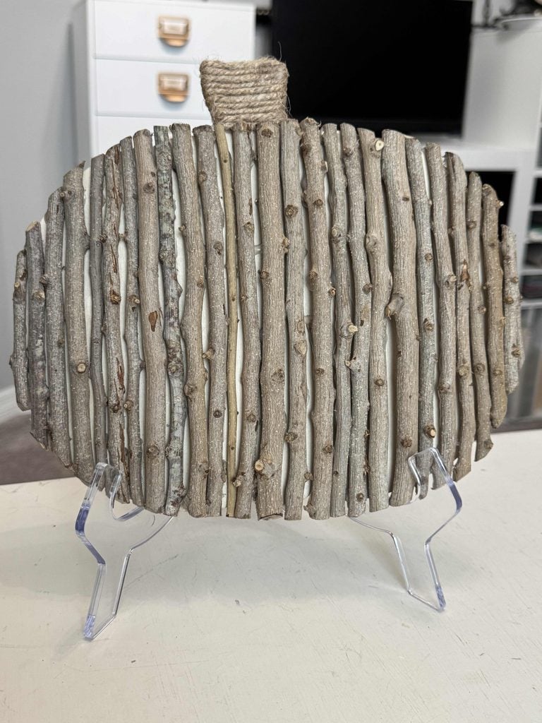 A charming DIY Twig Pumpkin features a pumpkin shape crafted from vertical wooden sticks, with a twine-wrapped stem, displayed on a clear stand. White drawers and shelves are visible in the background.
