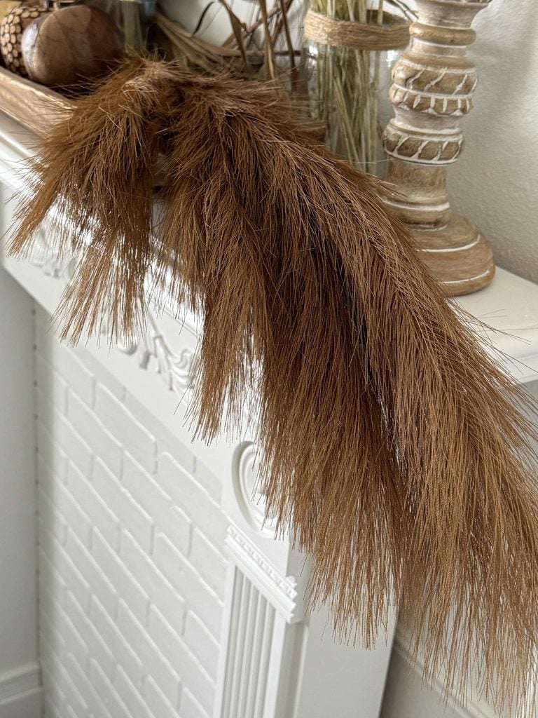 Brown dried pampas grass decor drapes over a white fireplace mantel, with decorative objects in neutral tones placed nearby, creating a natural, rustic aesthetic.
