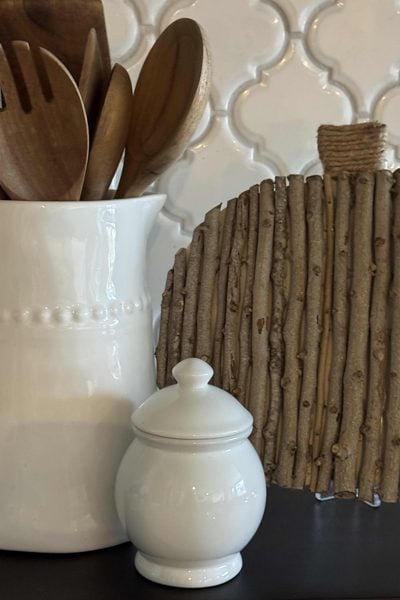 White ceramic jar and utensil holder with wooden spoons beside a charming DIY Twig Pumpkin, crafted from vertical sticks. The background features a white, textured tile pattern.