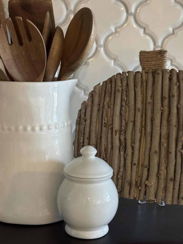 White ceramic jar and utensil holder with wooden spoons beside a charming DIY Twig Pumpkin, crafted from vertical sticks. The background features a white, textured tile pattern.