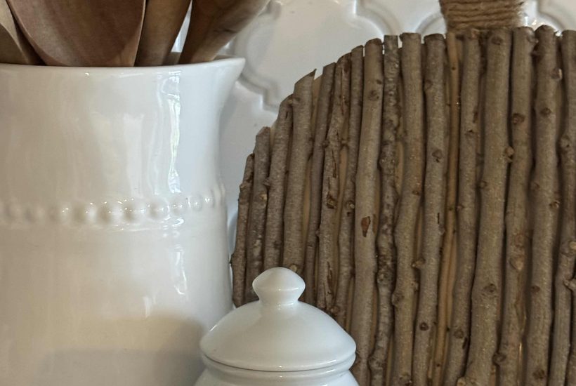 White ceramic jar and utensil holder with wooden spoons beside a charming DIY Twig Pumpkin, crafted from vertical sticks. The background features a white, textured tile pattern.