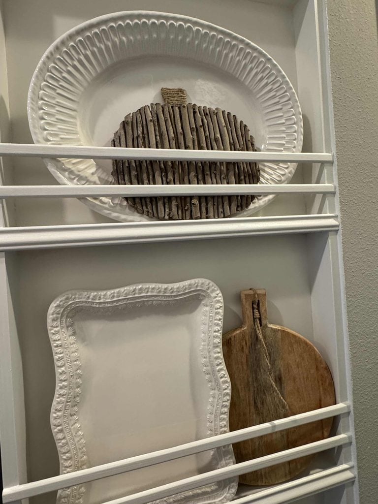 White plate rack with a decorative plate featuring a wood stick pumpkin—perfect for showcasing your favorite DIY pumpkin ideas—a square white plate with a scalloped edge, and a round wooden cutting board with a handle.