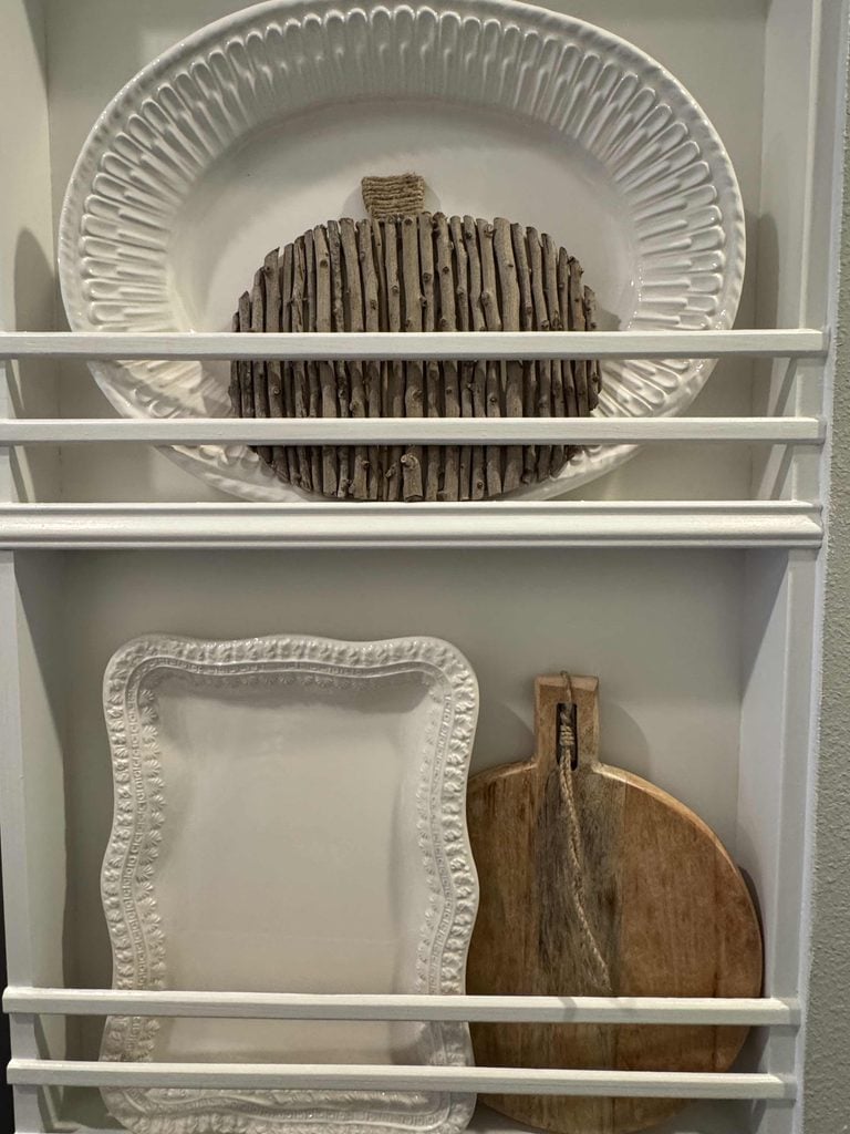 White shelves display decorative plates: a white platter, a DIY Twig Pumpkin shaped like a plate made of sticks, a rectangular white plate with a textured border, and a round wooden cutting board with a handle.