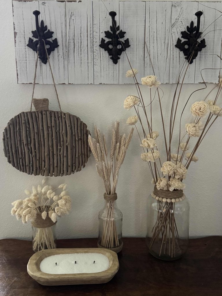 Three glass jars hold dried flowers and wheat stems on a wooden table. A rustic wood pumpkin, paired with a charming DIY Twig Pumpkin, and decorative hooks hang on a white wall above. A wooden tray with a white candle sits in the foreground.