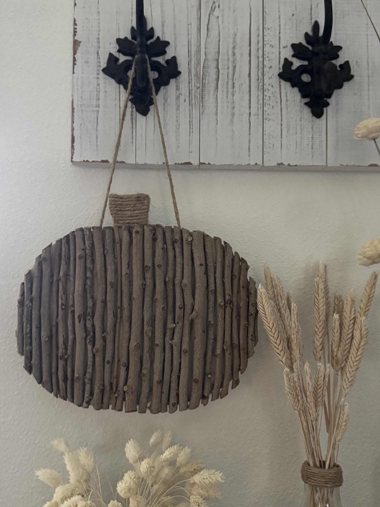 A rustic DIY Twig Pumpkin wall hanging, shaped like a pumpkin and crafted from vertical sticks with twine, is displayed on a white wall near dried wheat and grass in neutral tones.