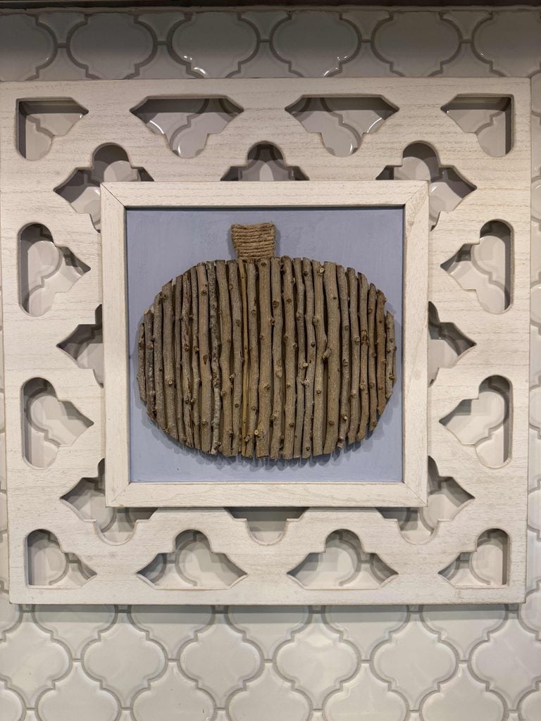 A wall decoration featuring a DIY Twig Pumpkin, crafted from vertical sticks arranged in a pumpkin shape on a blue background, framed by white wood with decorative cutouts, hanging on a tiled wall.