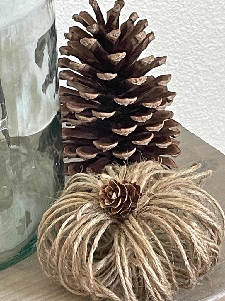 A large pinecone stands behind a rustic pumpkin decoration made of twine with a small pinecone on top, all placed on a wooden surface next to a glass jar.