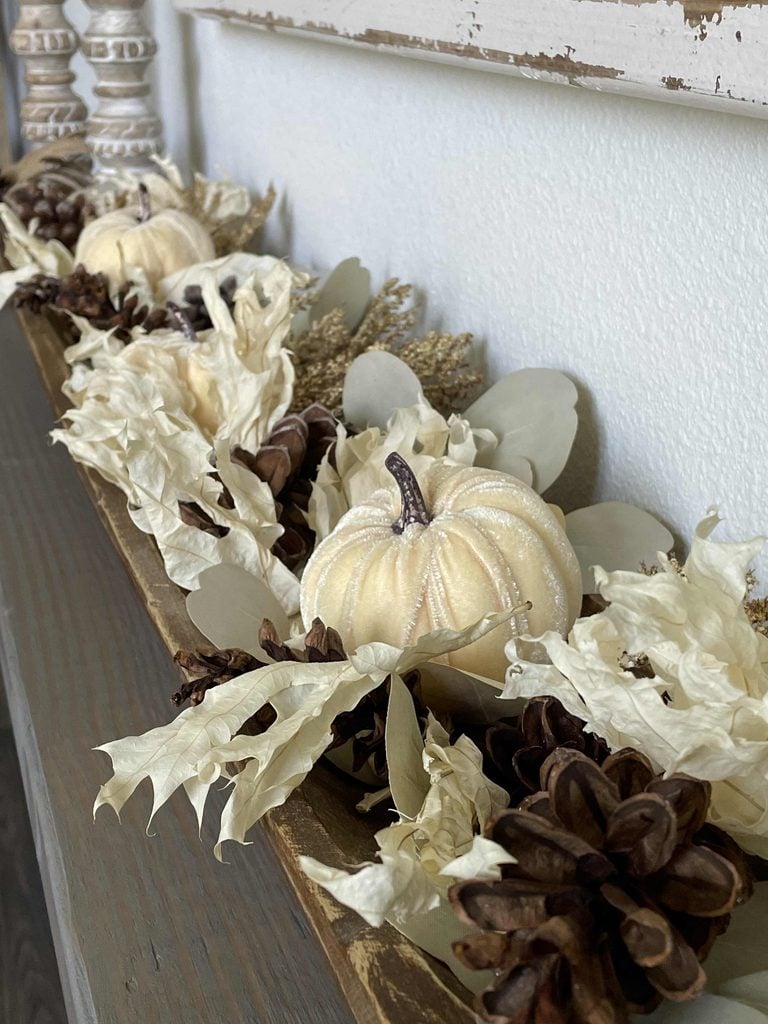 A wooden tray decorated with white pumpkins, pinecones, dried leaves, and foliage sits on a wooden surface against a white wall, creating a rustic autumn centerpiece.