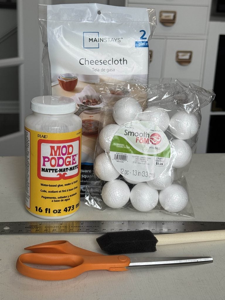Craft supplies on a table: cheesecloth, a bag of foam balls, a bottle of Mod Podge glue, orange scissors, a black foam brush, and a ruler. White drawers are in the background.