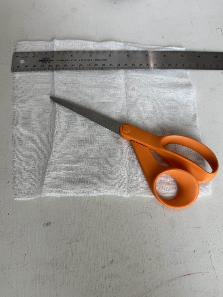 Orange scissors rest on white gauze with a metal ruler above, all placed on a white surface. The items suggest preparation for cutting the gauze to a specific size.