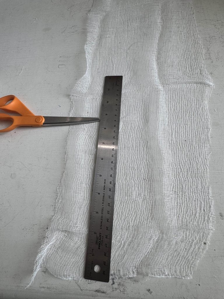 A pair of orange scissors and a metal ruler are placed on a piece of white gauze fabric lying on a light gray surface.