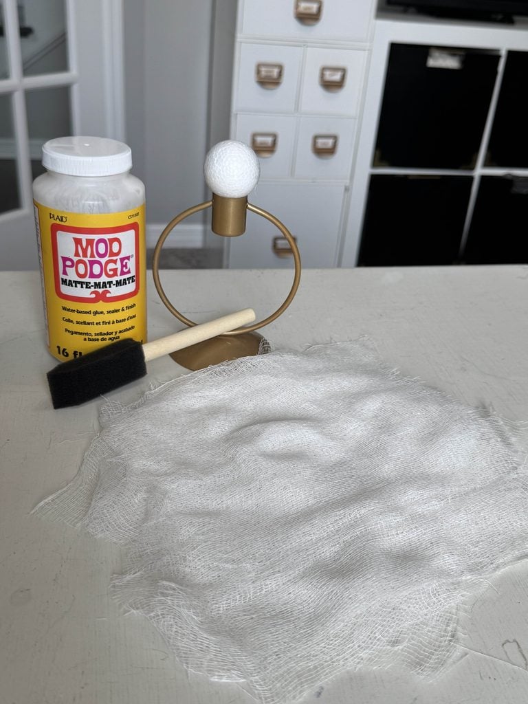 A container of Mod Podge, a foam brush, a metal stand with a styrofoam ball, and a piece of white gauze cloth are placed on a light gray table with storage cubbies in the background.
