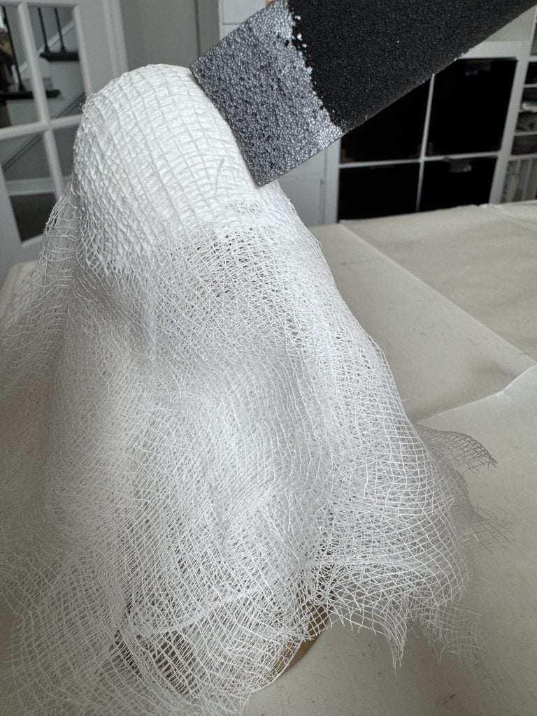White gauze draped over an object, resembling a ghost, with a paintbrush applying gray paint to the top; background shows a white table and shelves.