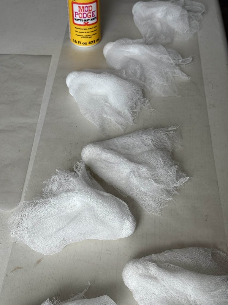 Six white gauze masks lie on a table next to a bottle of Mod Podge glue. The masks appear to be handmade and are textured with frayed edges.
