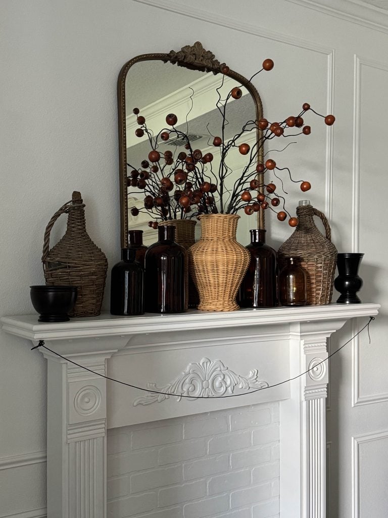 A white mantel with brown glass bottles, wicker vases, and dried branches. A large mirror reflects the room. Two black bowls sit at each end. The mantel is detailed and set against a white paneled wall.